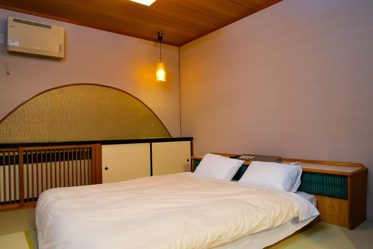 A hotel room with a bed, a wooden headboard, a wall-mounted air conditioner, and a hanging light fixture.