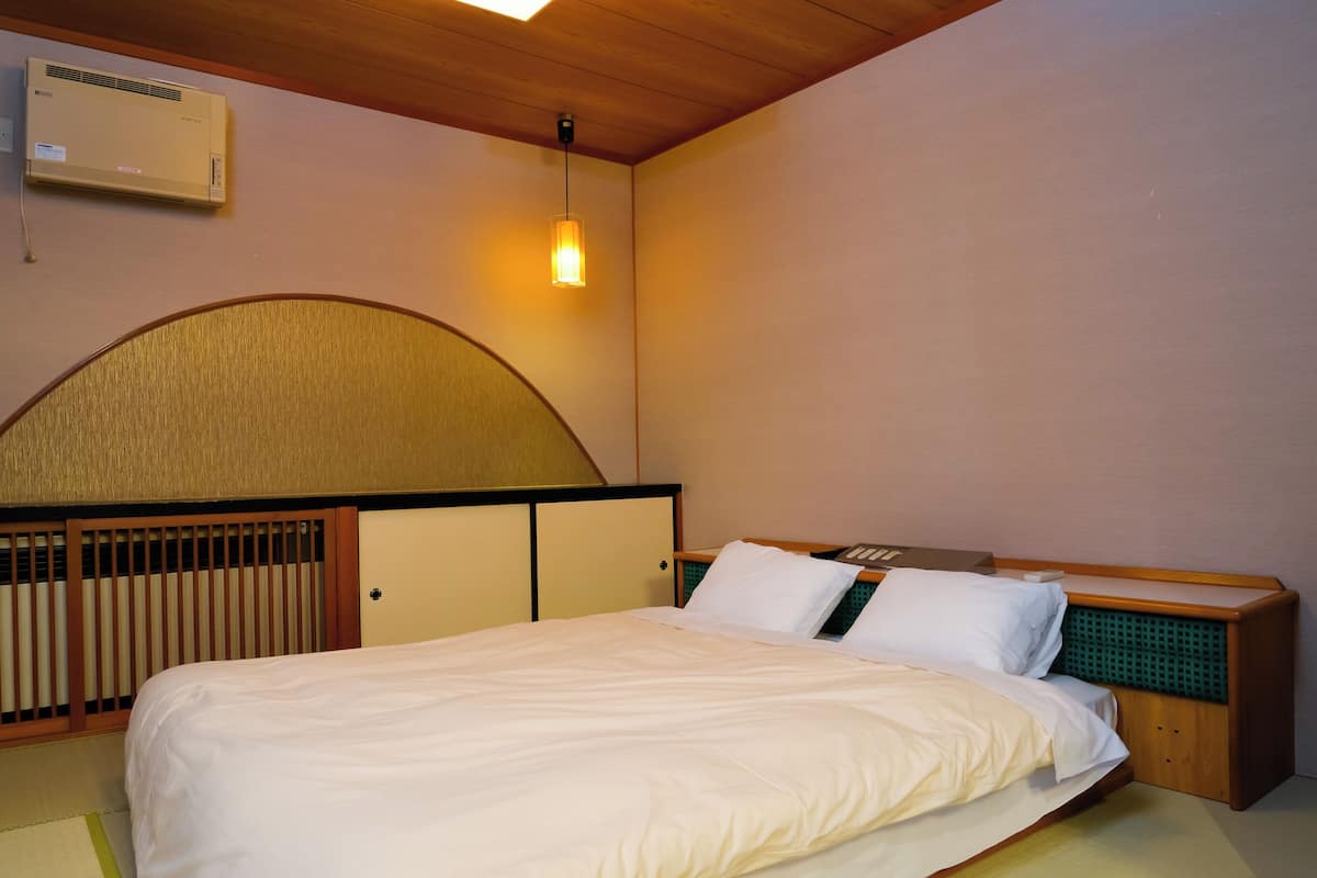 A hotel room with a bed, a bedside table, a lamp, and an air conditioning unit.