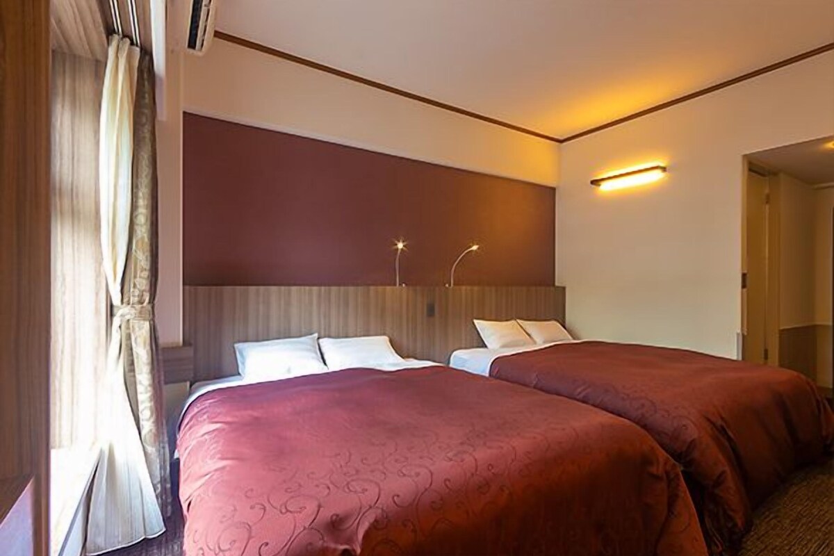 A hotel room with two beds, a wooden headboard, a wall-mounted light, and a window with curtains.