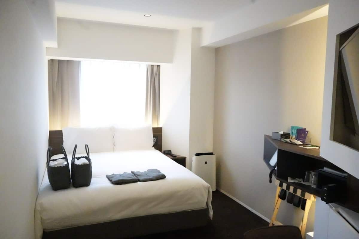 Une chambre d’hôtel avec un lit, un bureau avec un ordinateur, une télévision et une fenêtre avec des rideaux.