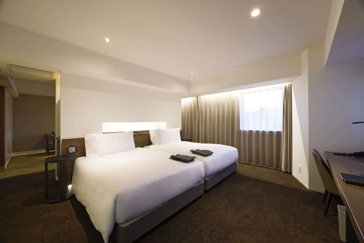 Une chambre d’hôtel avec un grand lit, un bureau, une chaise, une lampe et une fenêtre avec des rideaux.