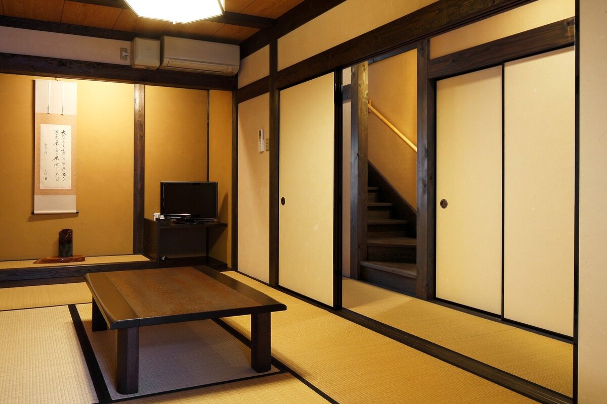 Une pièce de style japonais traditionnel, avec un sol recouvert de tatamis, une table basse, un téléviseur, des portes coulissantes et un escalier.