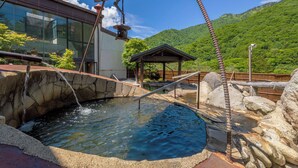 Aguas termales con paredes de piedra, una estructura de madera y montañas al fondo.