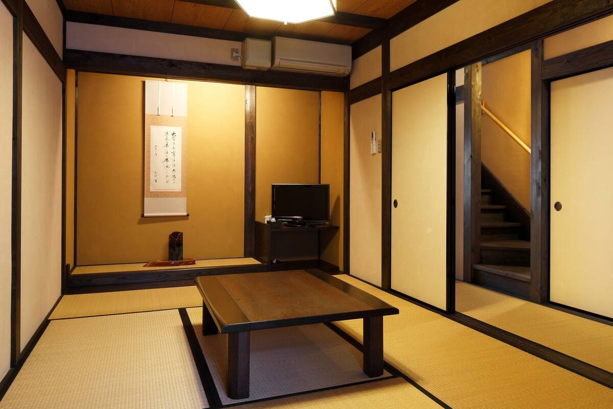 Una habitación japonesa tradicional con suelo de tatami, una mesa baja, televisor, puertas correderas y una escalera.
