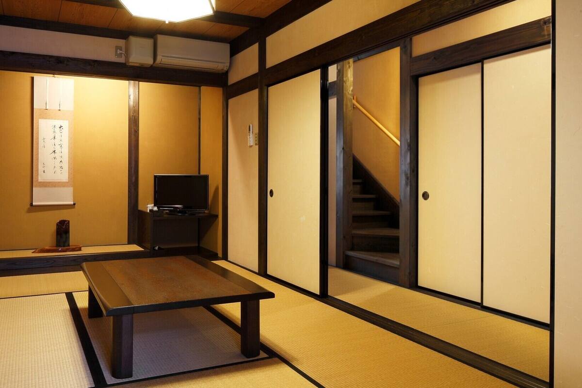 Una habitación tradicional japonesa con suelo de tatami, una mesa baja, televisor, puertas correderas y una escalera.