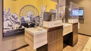 A hotel reception desk with a clear glass partition, a digital screen displaying the hotel name, and a wall mural featuring iconic landmarks. - Premier Inn Glasgow City Centre Hotel (Glasgow)
