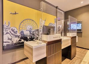 A hotel reception desk with a clear glass partition, a digital screen displaying the hotel name, and a wall mural featuring iconic landmarks.