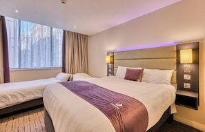 A hotel room with two beds, a large window with curtains, a headboard with integrated lighting, and a bedside table with a lamp.