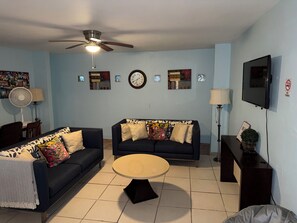 A living room with two sofas, a coffee table, a ceiling fan, wall clocks, and a TV.