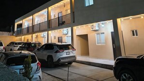 Un edificio de varios pisos con balcones, unidades de aire acondicionado y un estacionamiento con varios coches.