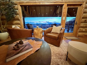 A cozy cabin interior with a leather sofa, a round table, and a view of mountains through a large window.
