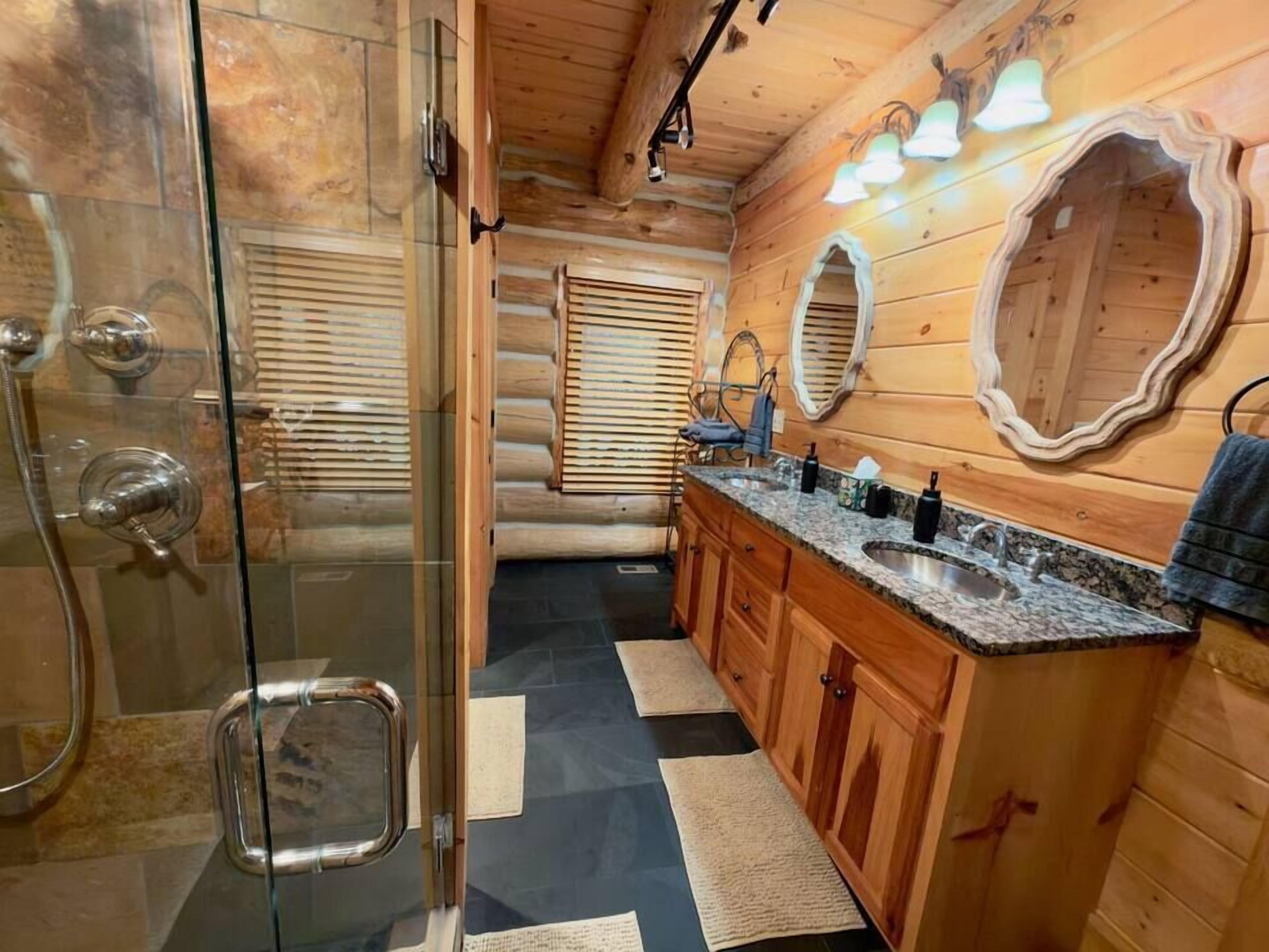 Une salle de bain rustique, aménagée dans une cabane en rondins, avec une douche à parois de verre, des meubles en bois, un comptoir en granit et un grand miroir.