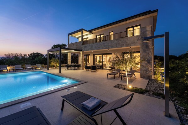 Une maison moderne à deux étages, dotée d’une piscine, d’un espace extérieur aménagé et d’un patio bien éclairé.