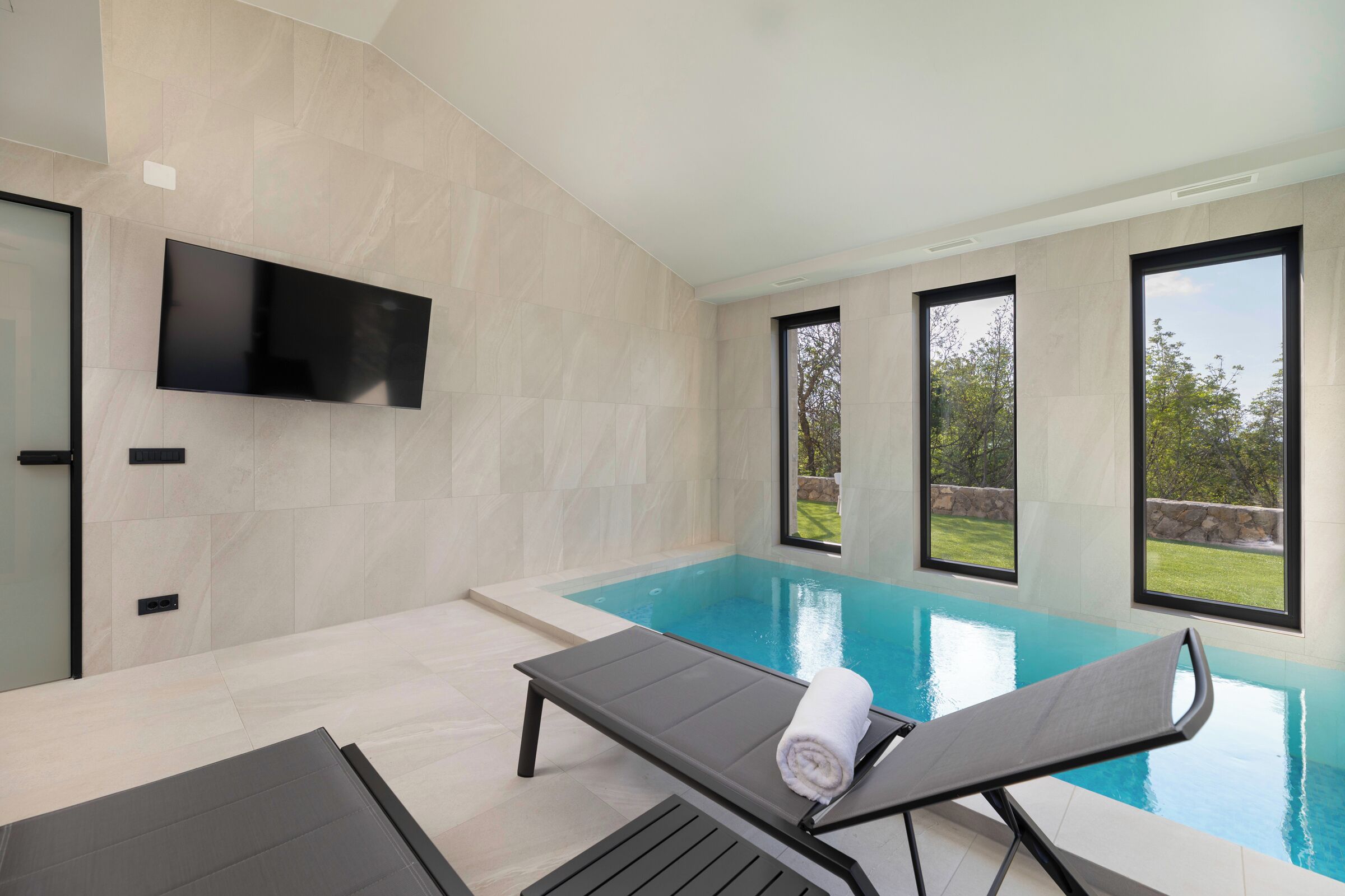 Una piscina interior moderna con techo plano, un televisor de pantalla plana montado en la pared y grandes ventanales con vistas al exterior.