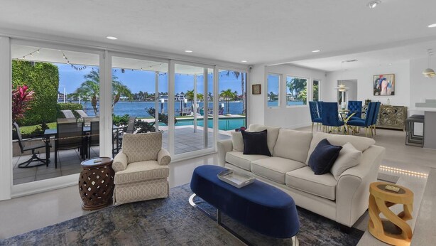 Uma sala de estar moderna com uma janela grande que dá para uma piscina e oceano, um sofá, uma mesa de centro e uma área de jantar com cadeiras azuis.