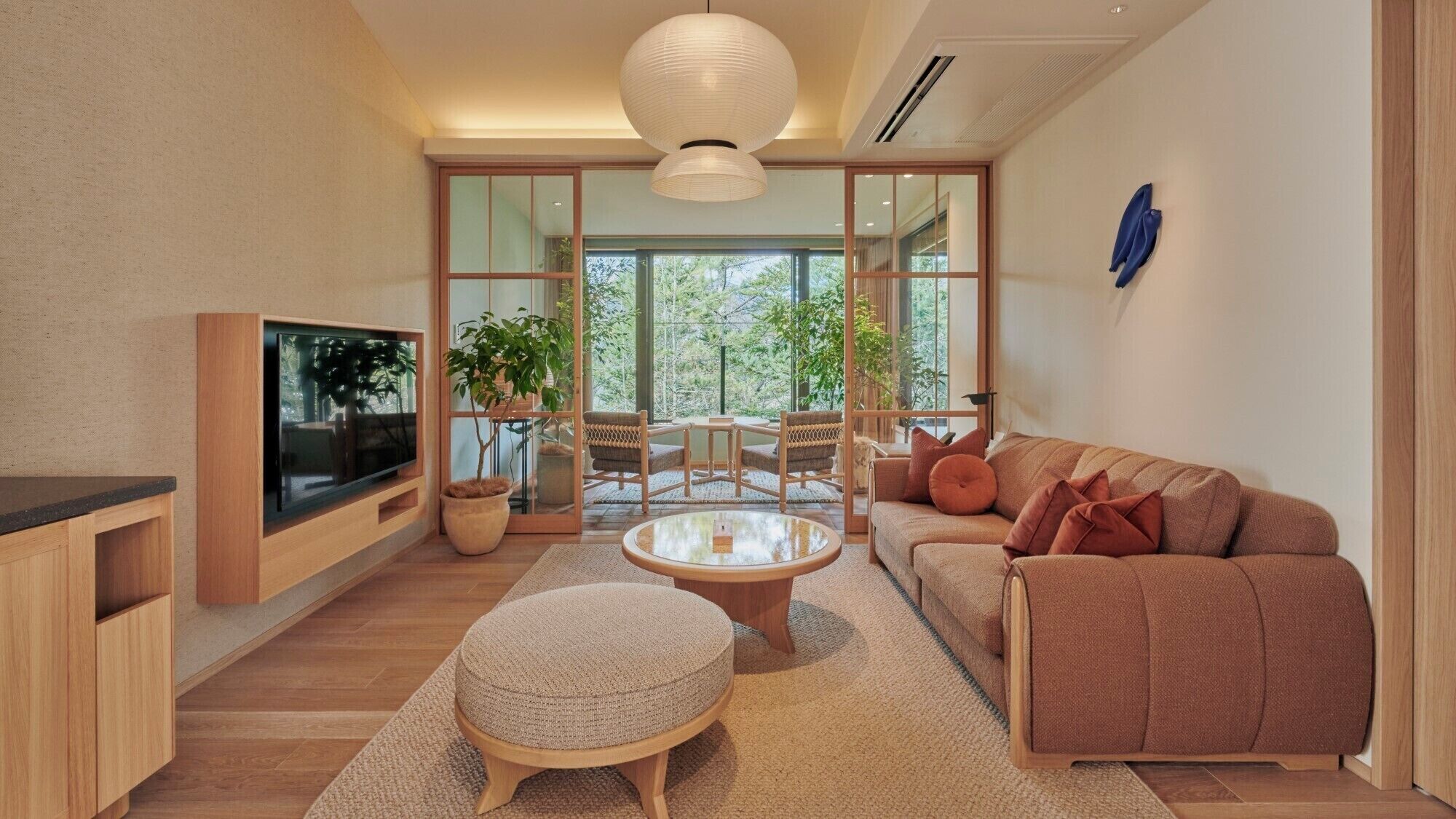 Un salon moderne avec un téléviseur, un canapé, un pouf et une grande fenêtre donnant sur la nature et les arbres.
