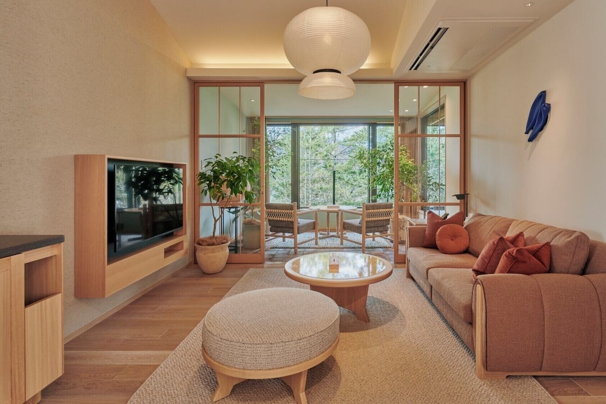 Uma sala de estar moderna com televisão, sofá, pufe e uma grande janela com vista para árvores.
