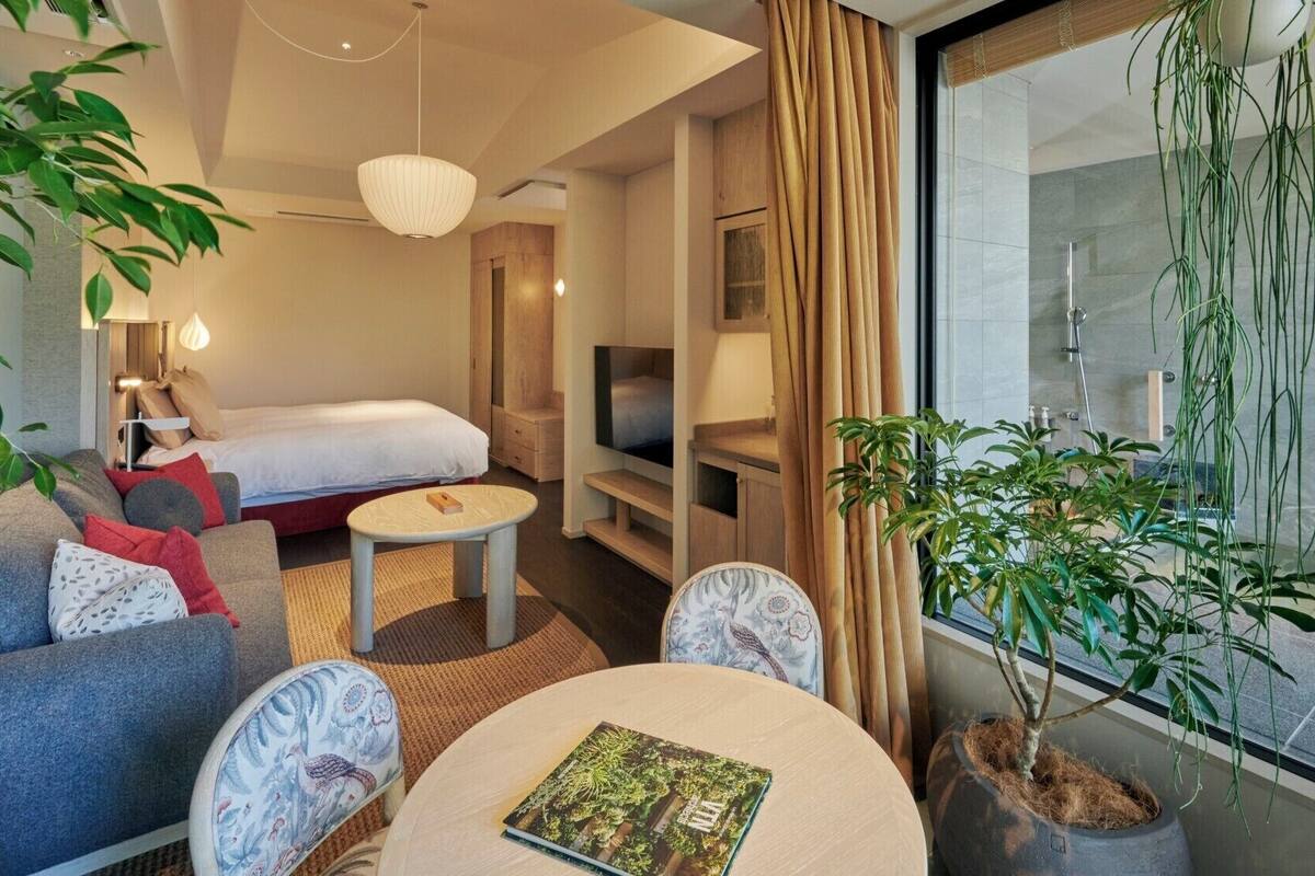 Quarto de hotel moderno com cama, sofá, TV e plantas em vasos.