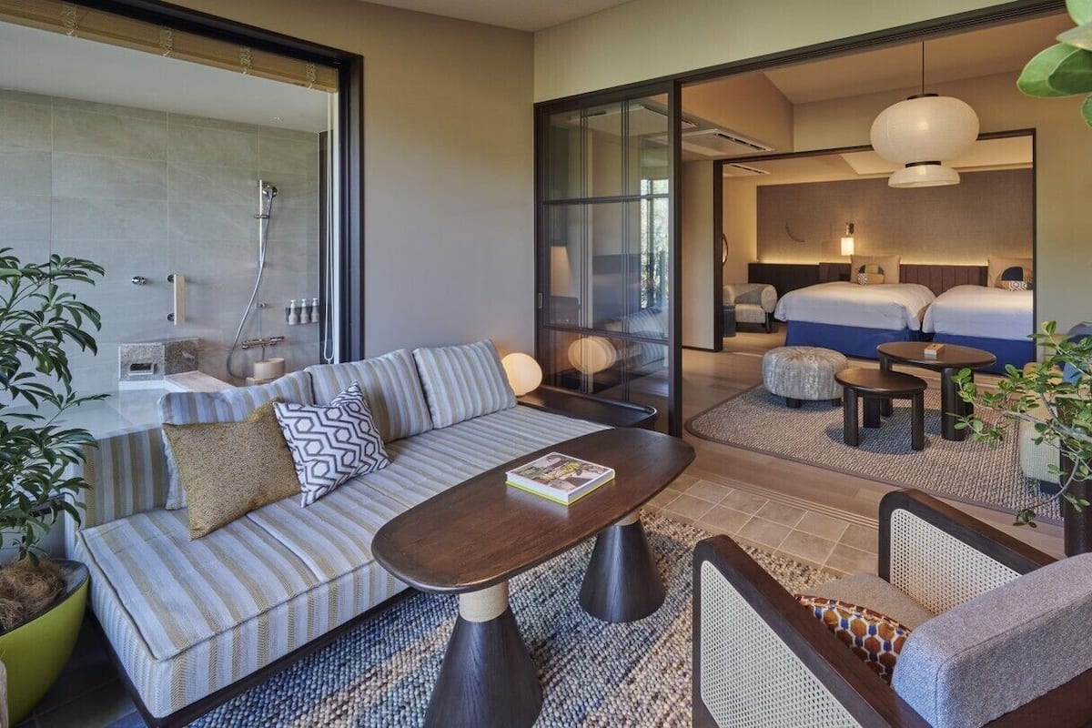 Quarto de hotel moderno com sofá, mesa de centro, cama com duas almofadas, mesa de cabeceira e uma janela grande.