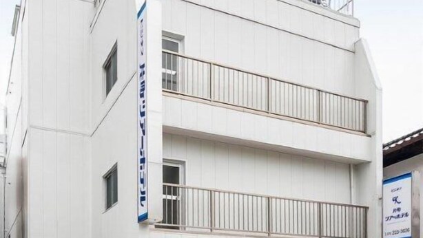 Ein mehrstöckiges Gebäude mit weißer Fassade, einem sichtbaren Eingang und einem Balkon im oberen Stockwerk.