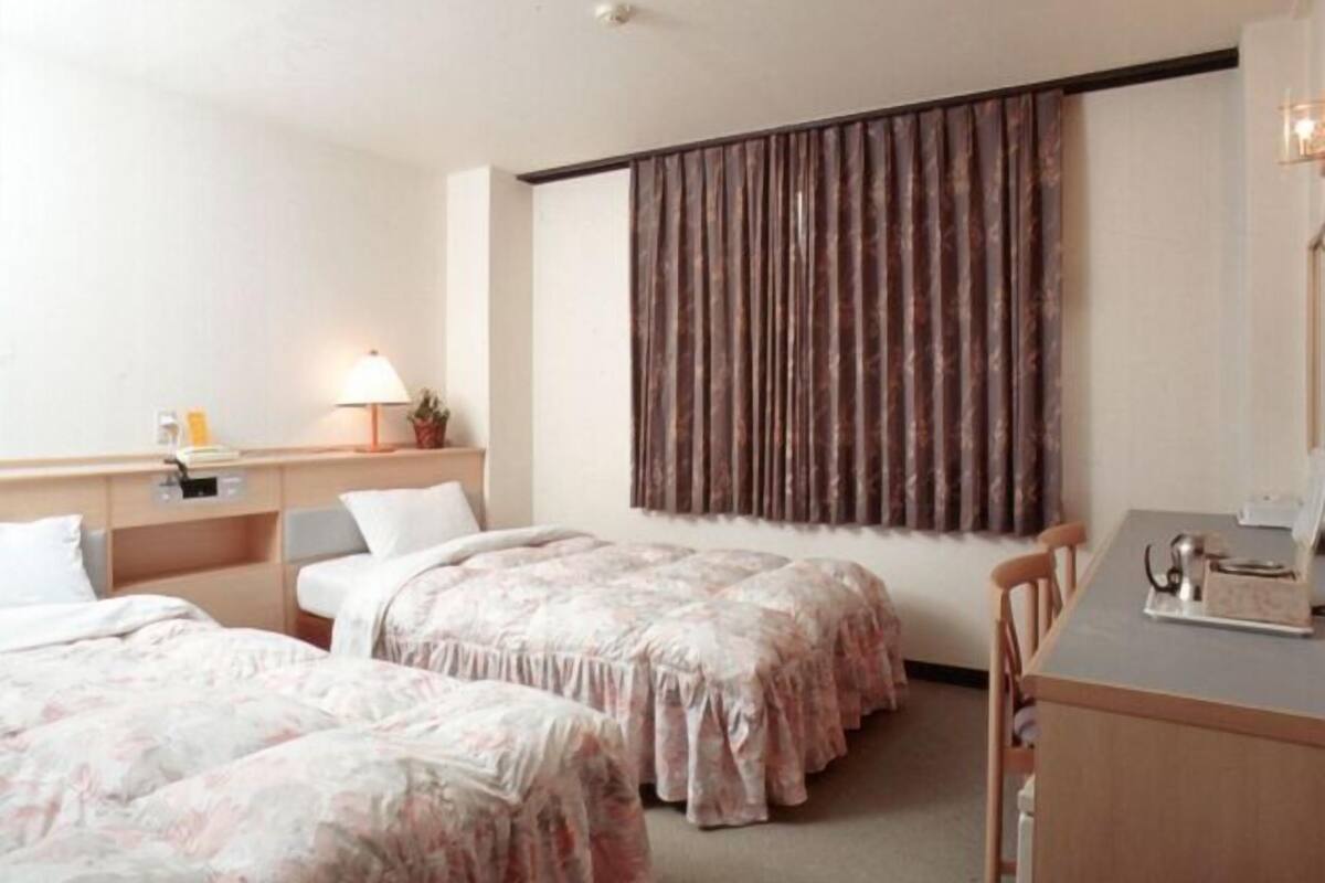 Habitación de hotel con dos camas, un escritorio, una silla, una lámpara y un ventanal grande con cortinas.