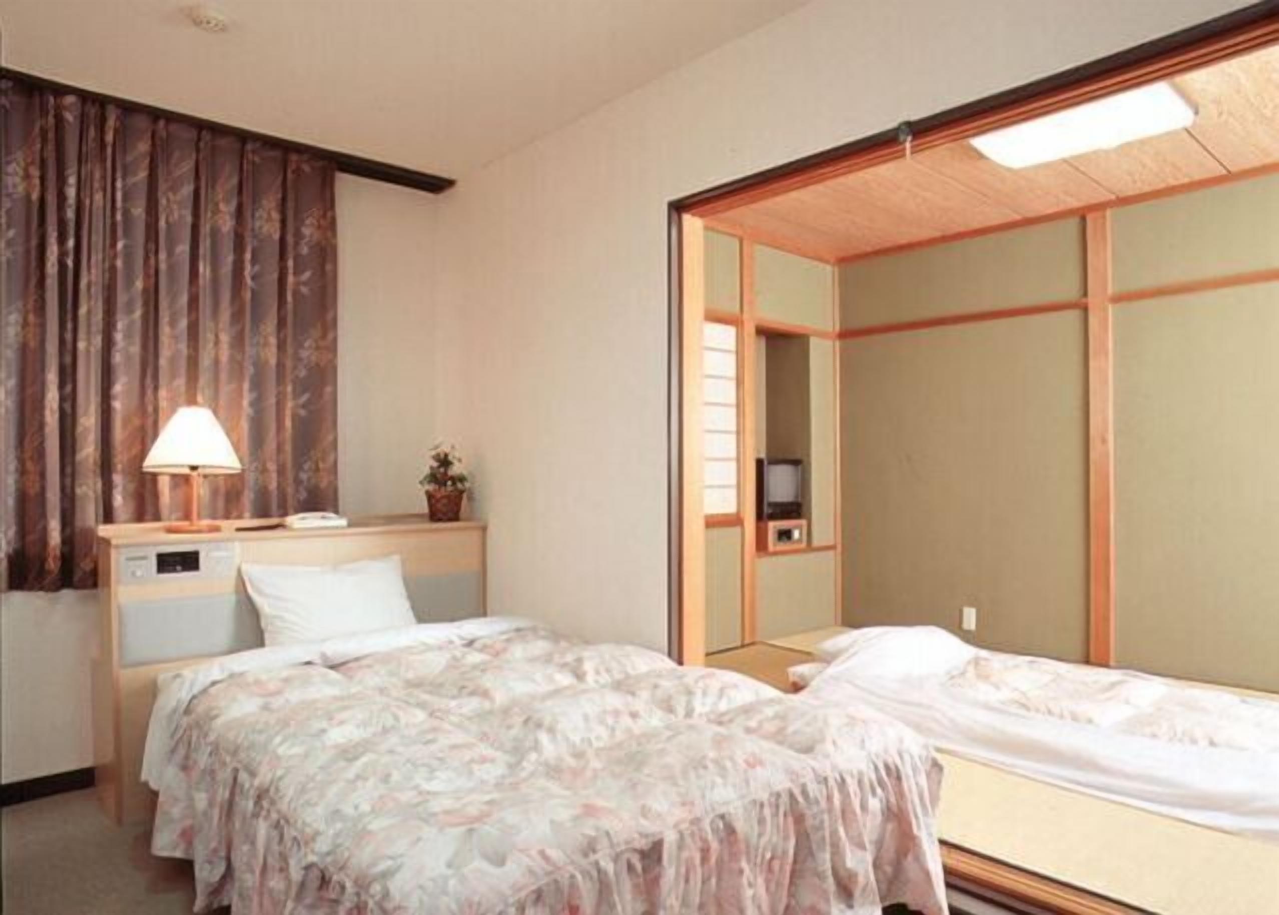 A hotel room with two beds, a bedside table with a lamp, a small plant, and a TV.