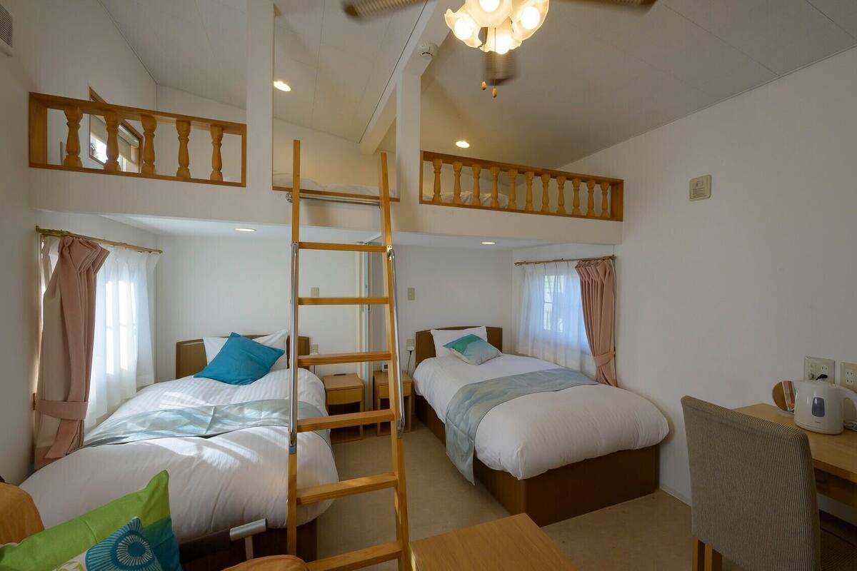 A room with two beds, a desk, a chair, a ladder, and a ceiling light fixture.