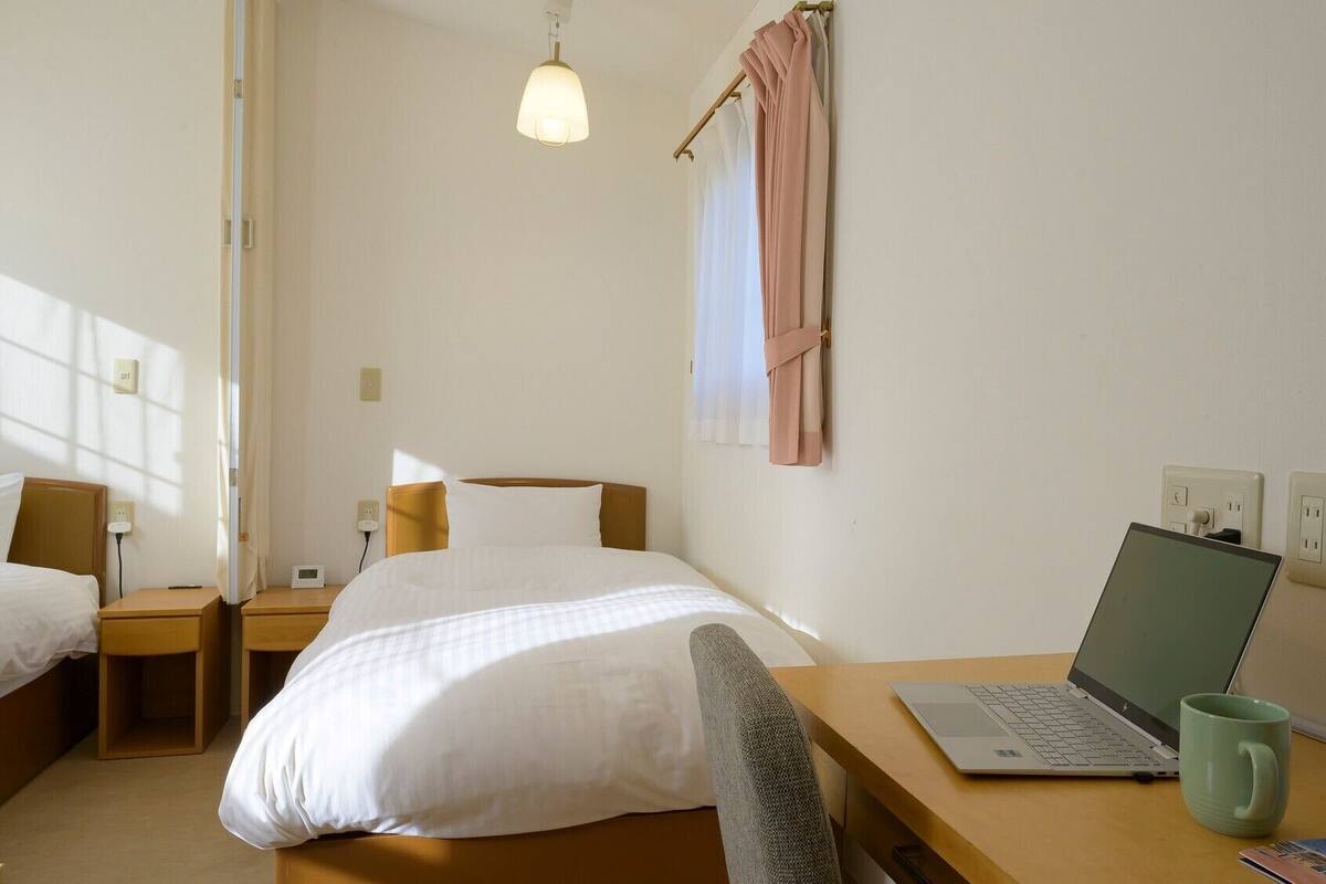 A hotel room with two beds, a desk with a laptop, a mug, and a window with curtains.
