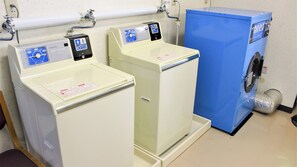 A coin washing machine and a blue laundry machine in a room with a white wall and a pipe above the machines. - Sounkyo Hostel (Kamikawa)