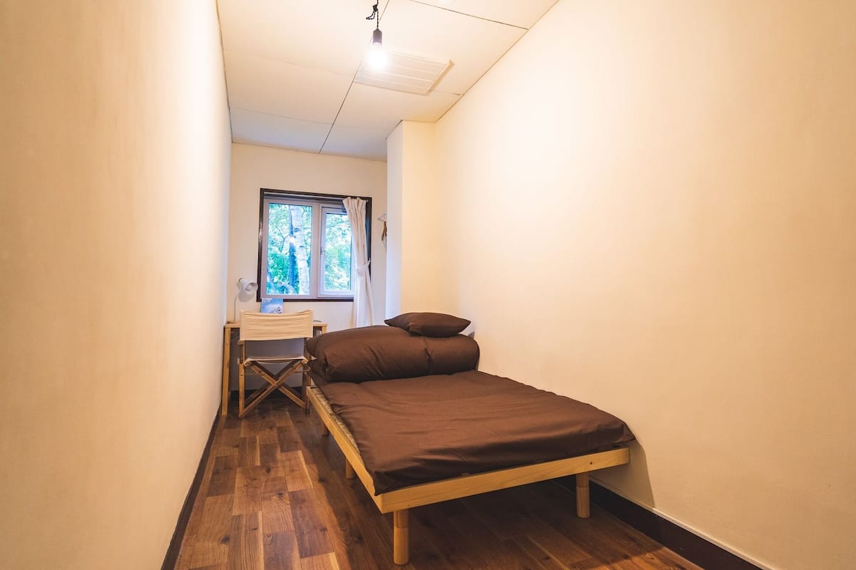 A small, narrow room with a bed, a desk, and a window.