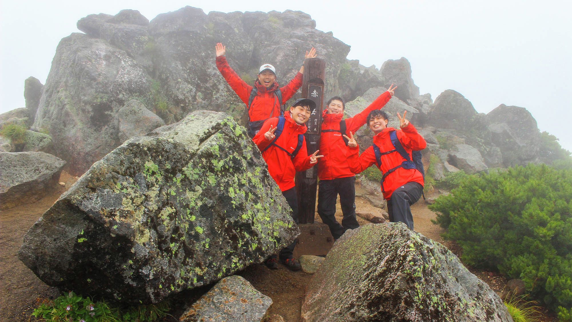 A group of people in red jackets posing on a rocky terrain with large boulders and greenery.