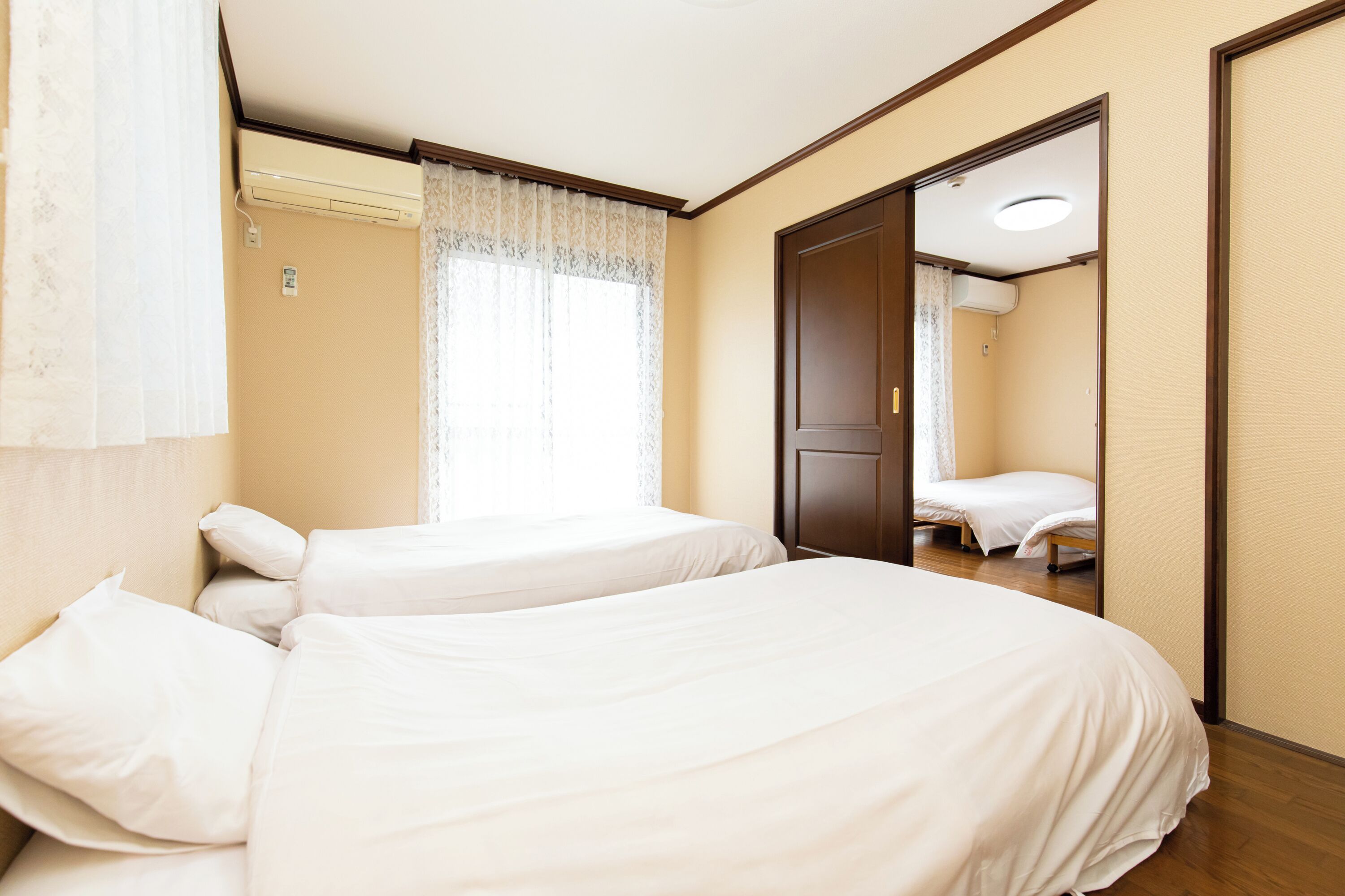 A hotel room with two beds, a window with curtains, an air conditioning unit, and a door leading to another room.