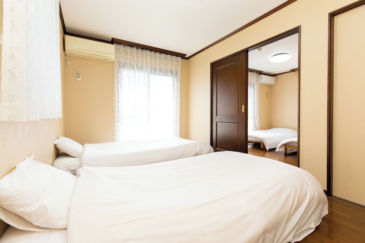 Ein Hotelzimmer mit zwei Betten, einem mit Vorhängen versehenen Fenster, einer Klimaanlage und einer Tür zu einem weiteren Raum.