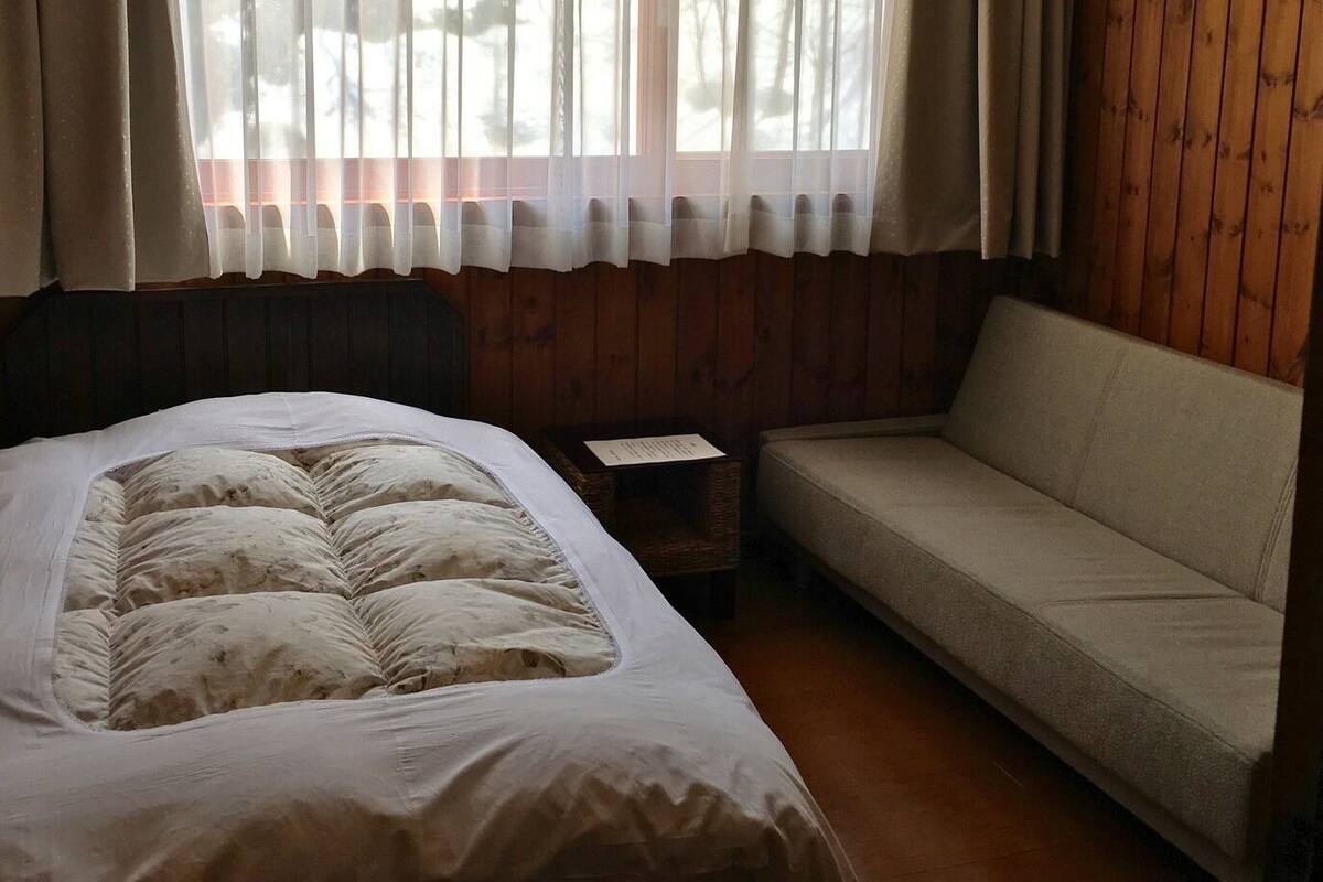 A bedroom with a bed, a sofa, a window with sheer curtains, and wooden paneling on the walls.