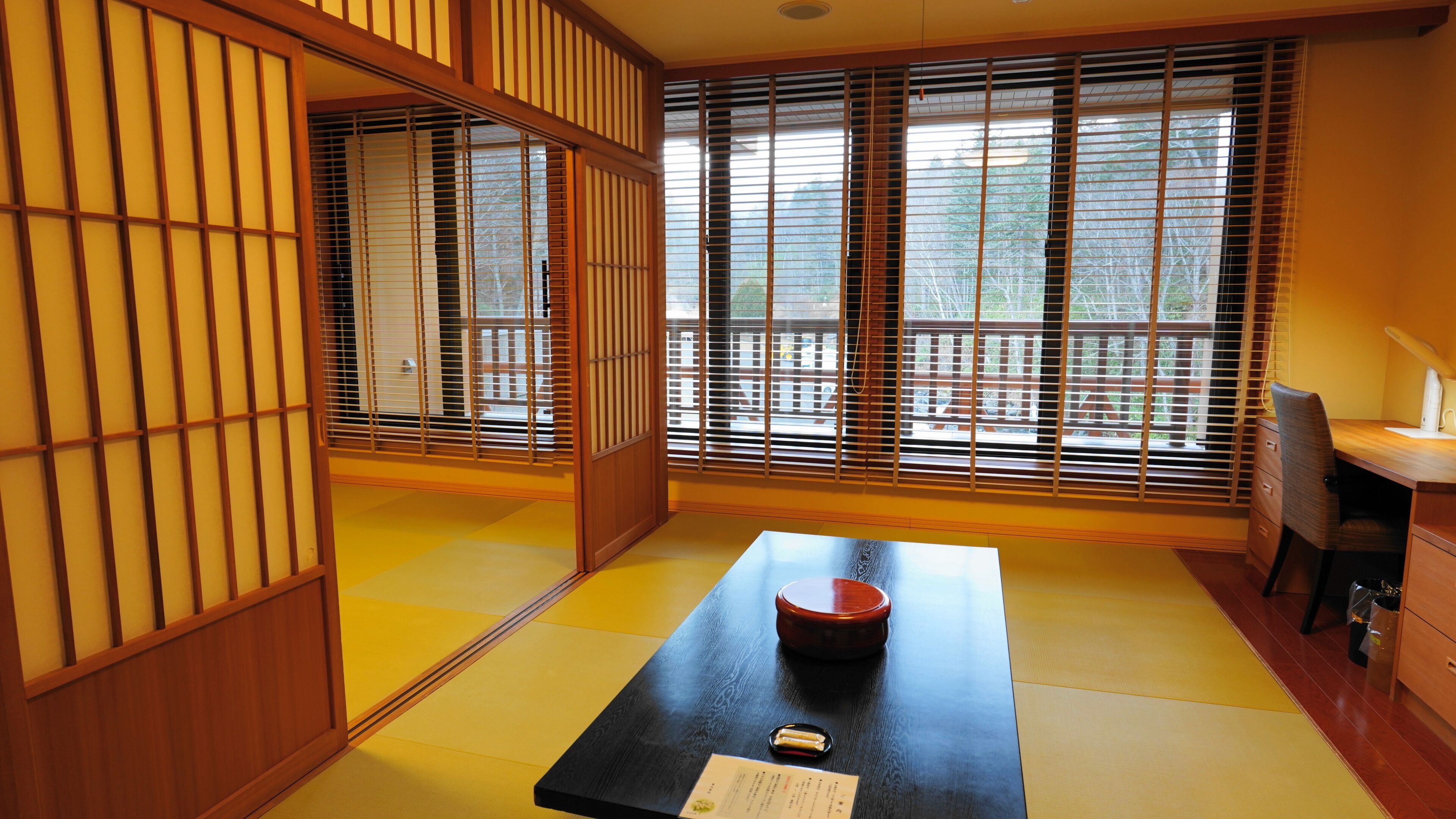 A traditional Japanese room with tatami flooring, shoji screens, a tatami mat table with a red tray, and a desk with a lamp.
