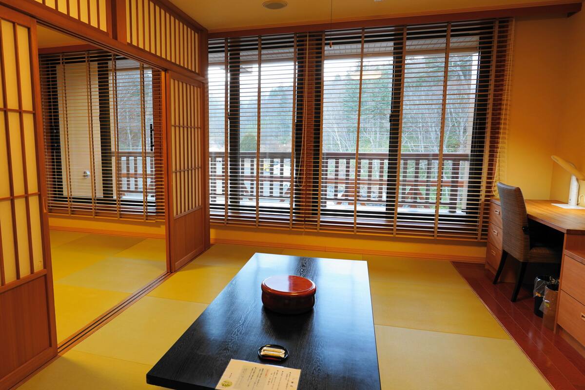 A traditional Japanese room with tatami flooring, shoji screens, a tatami mat table with a red tray, and a desk with a lamp.