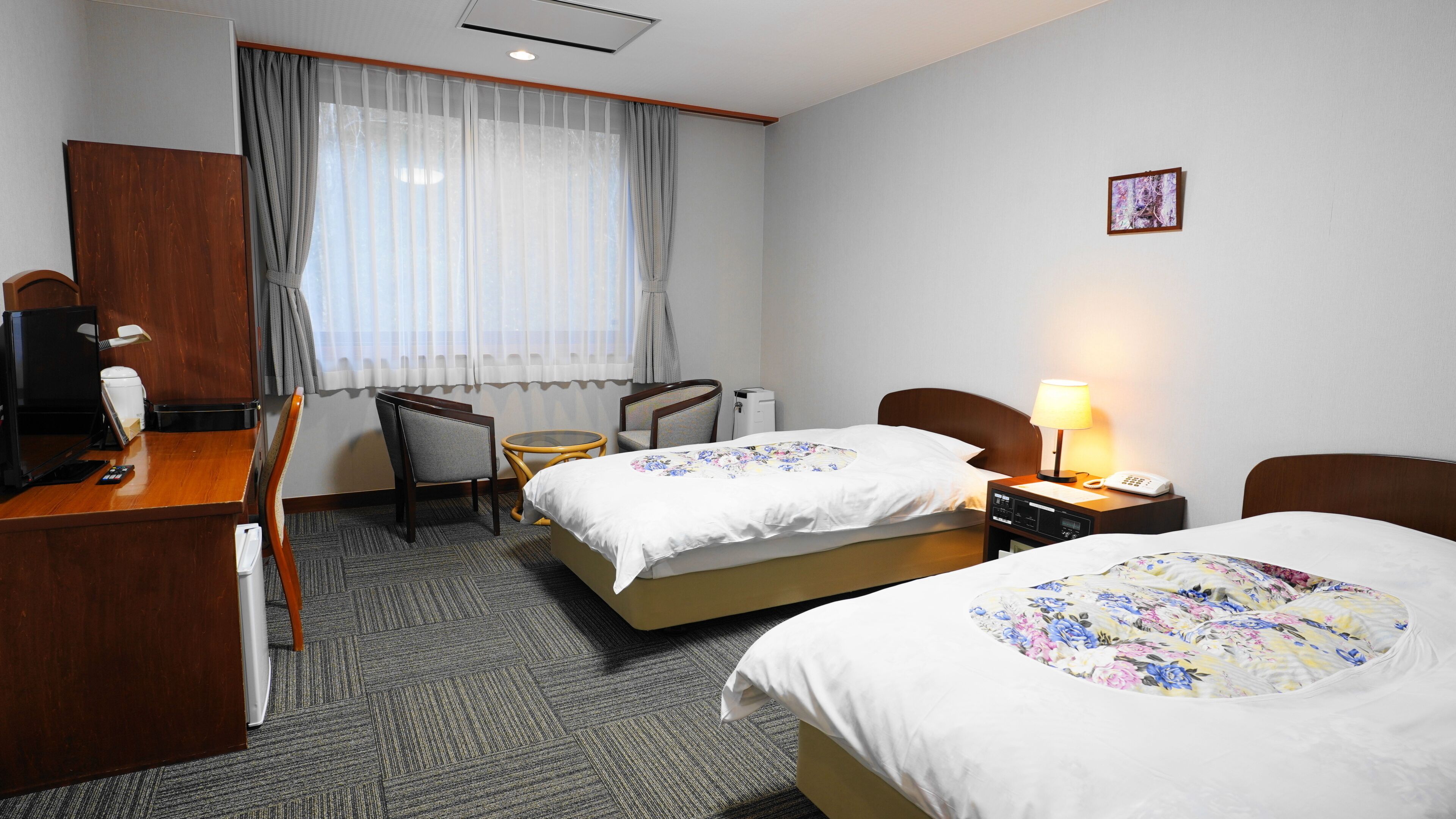 A hotel room with two beds, a desk, a chair, a small table, a lamp, and a picture on the wall.
