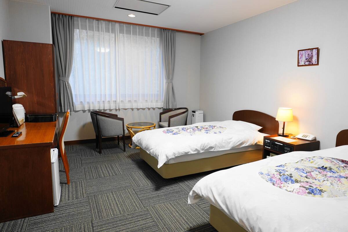 A hotel room with two beds, a desk, a chair, a small table, a lamp, and a picture on the wall.