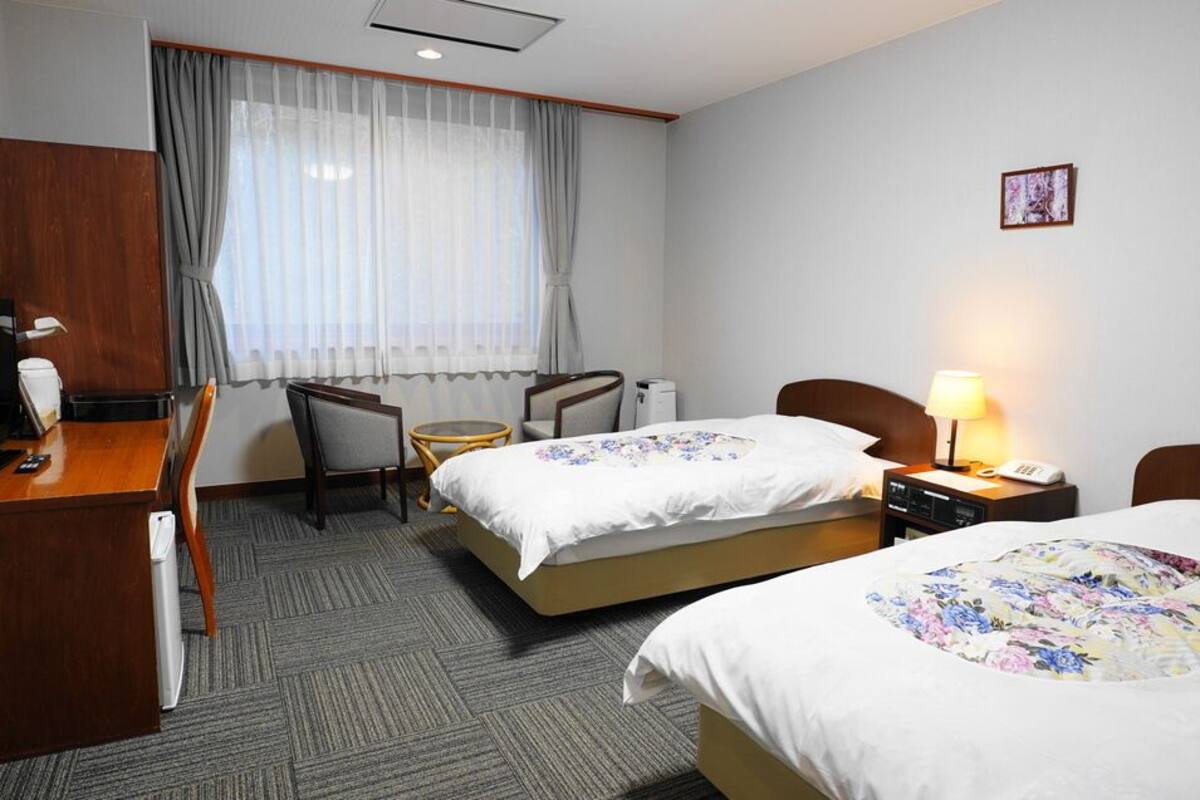 A hotel room with two beds, a desk, a chair, a small table, a lamp, and a window with curtains.