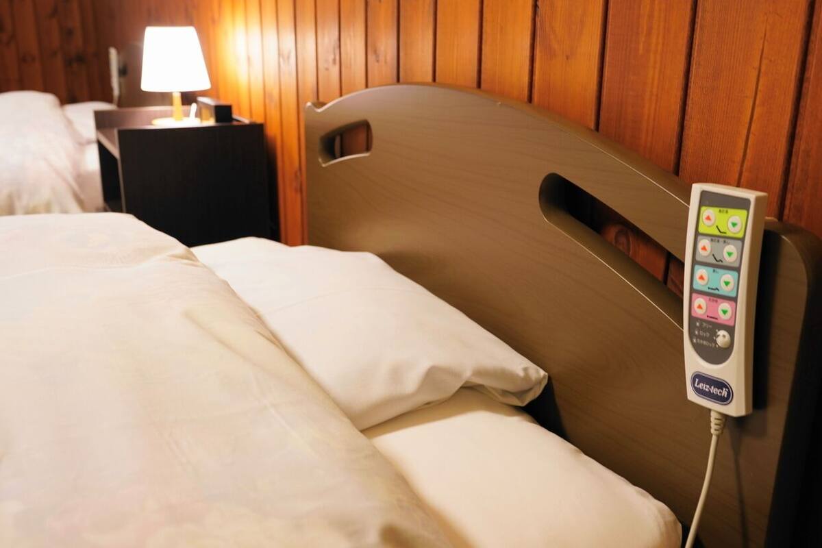 A bed with a control panel attached to the headboard, a bedside table with a lamp, and wooden paneling on the wall.