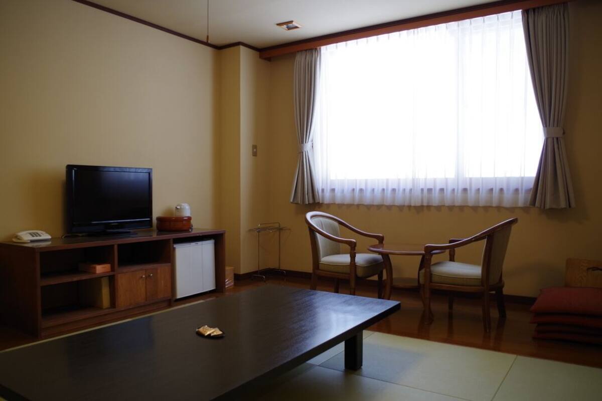 A room with a television on a wooden stand, a small cabinet, a table, two chairs, and a window with curtains.