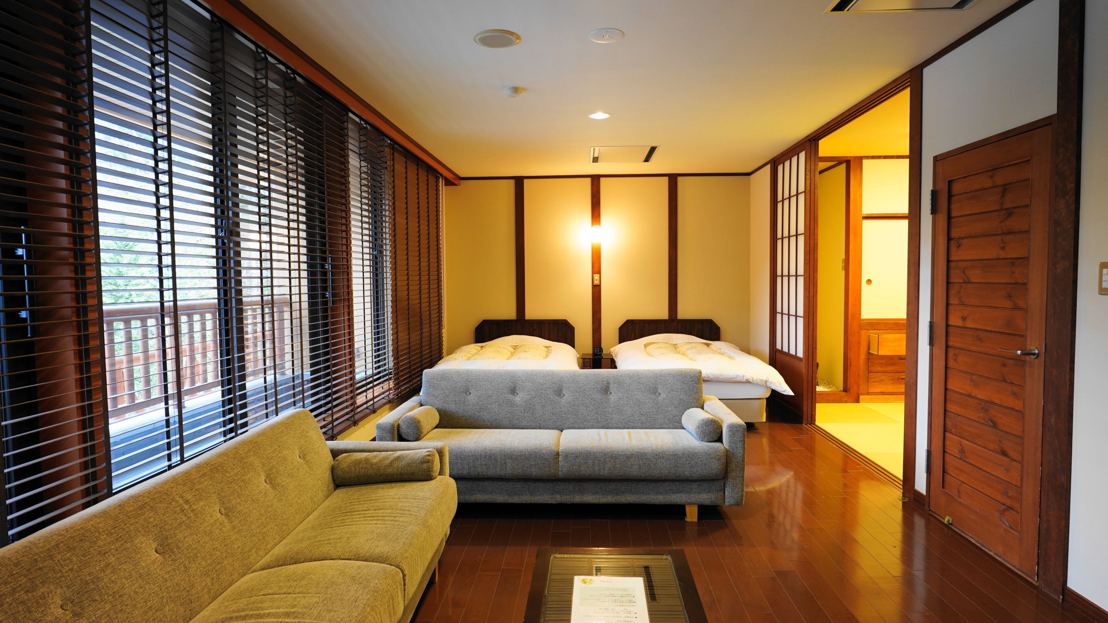 A hotel room with a sofa, two beds, wooden flooring, and a sliding door.
