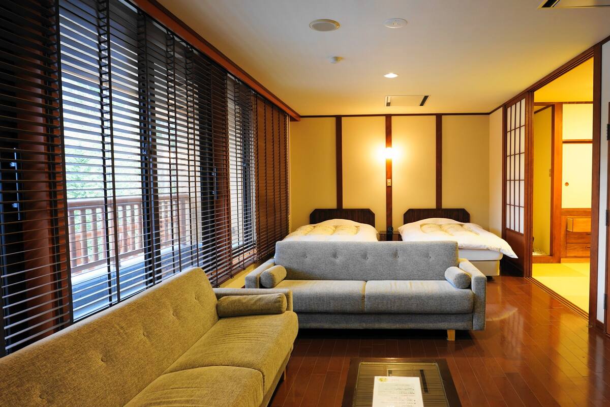A hotel room with a sofa, two beds, wooden flooring, and a sliding door.