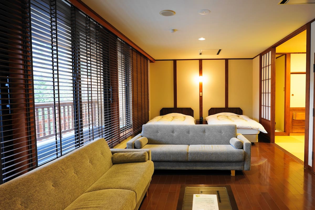 A hotel room with a sofa, two beds, wooden flooring, and a sliding door.