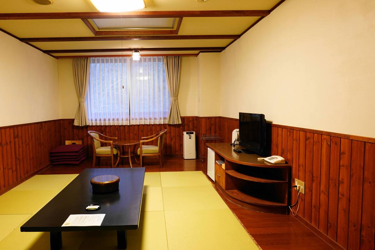 A room with a tatami mat floor, a low table with a placemat, a chair, a small cabinet, and a television on a wooden stand.