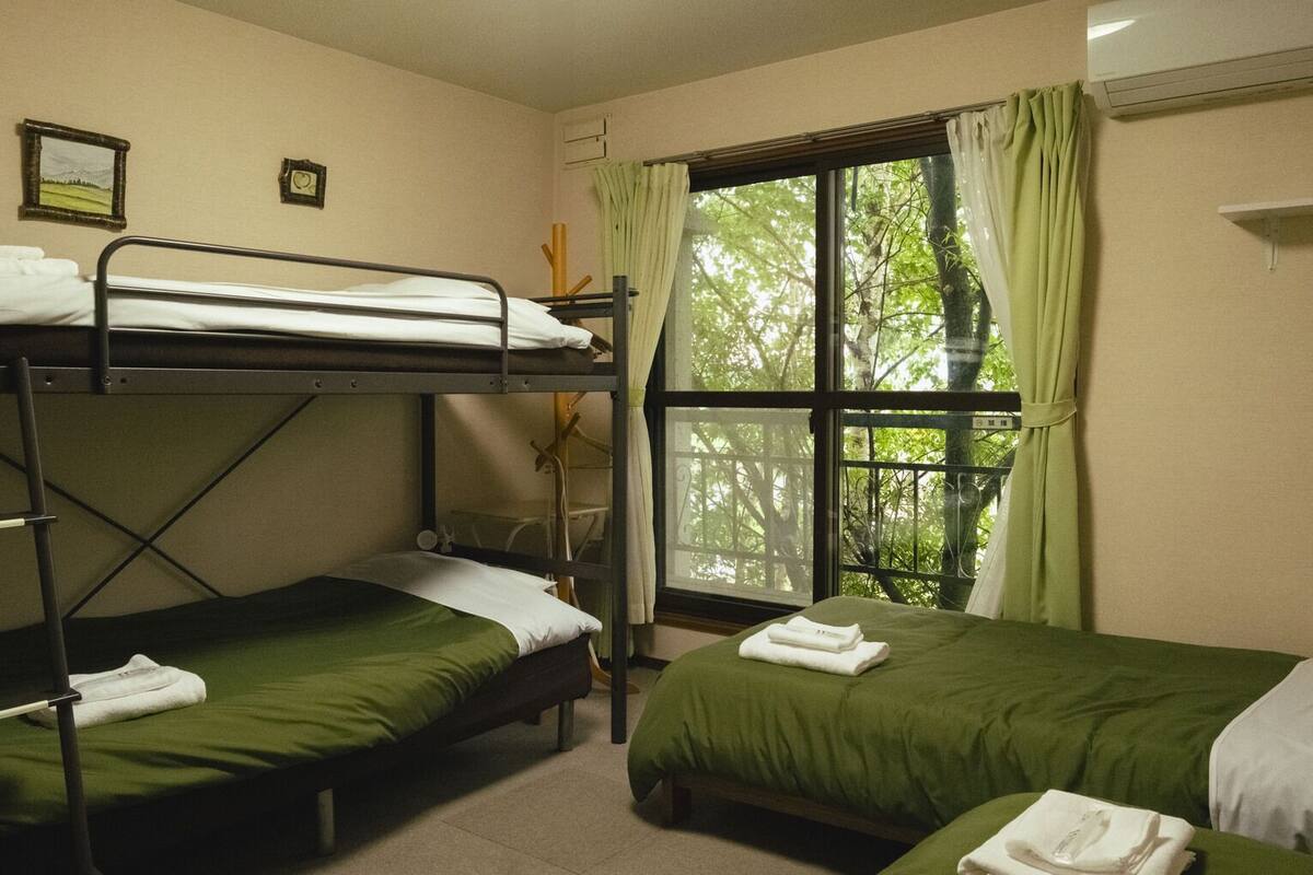 A dormitory room with bunk beds, a window with curtains, and a bed with a green bedspread.