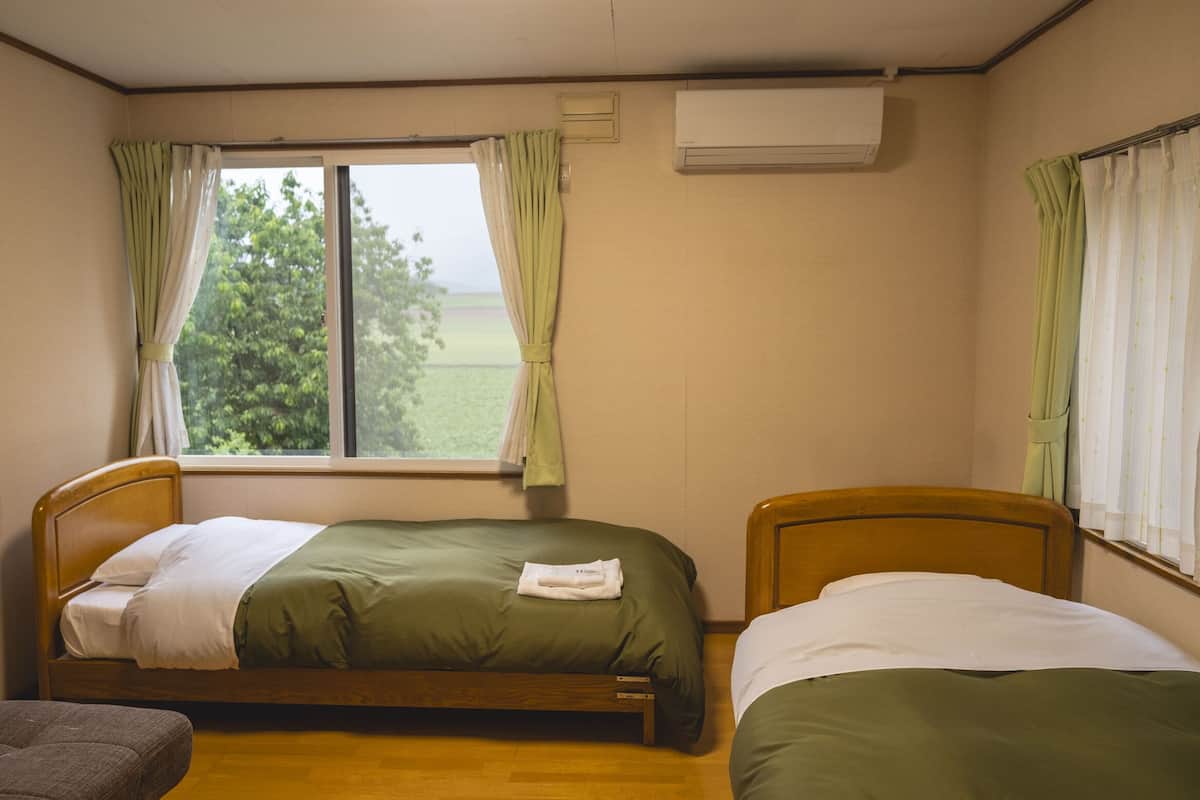 A room with two single beds, a wooden headboard, a window with curtains, and an air conditioning unit.