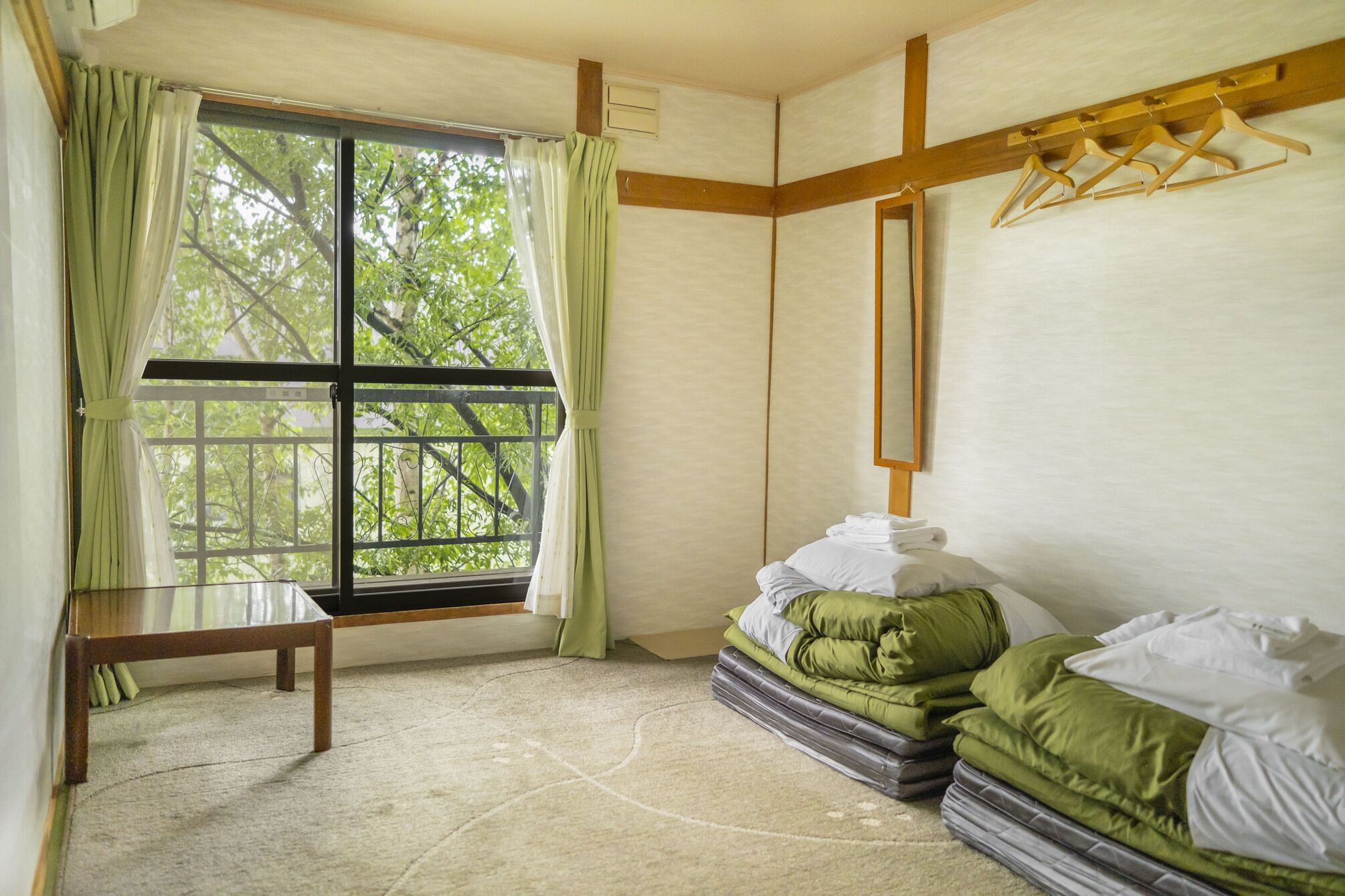 Een kamer met een bed, een bankje, een raam met gordijnen en een houten plank met kledinghangers.