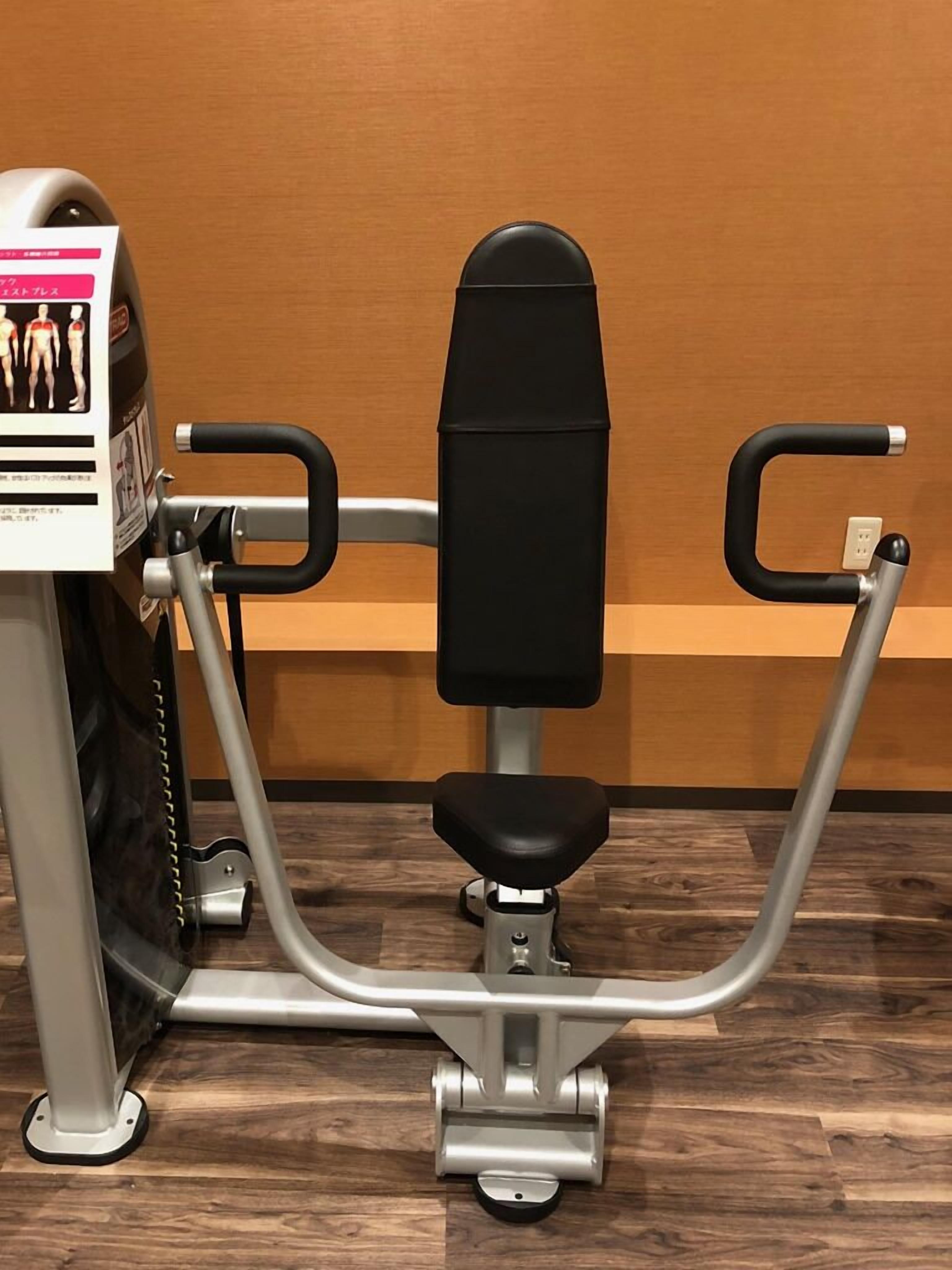 Een stilstaande fitnessmachine met een zwarte zitting en handgrepen, een bedieningspaneel met een display en een wandmontage basis.