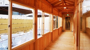 A corridor with wooden walls and large windows, leading to an outdoor area with tables and chairs. - Capsule Hotel Topos Sendai Station Nishiguchi (Sendai)